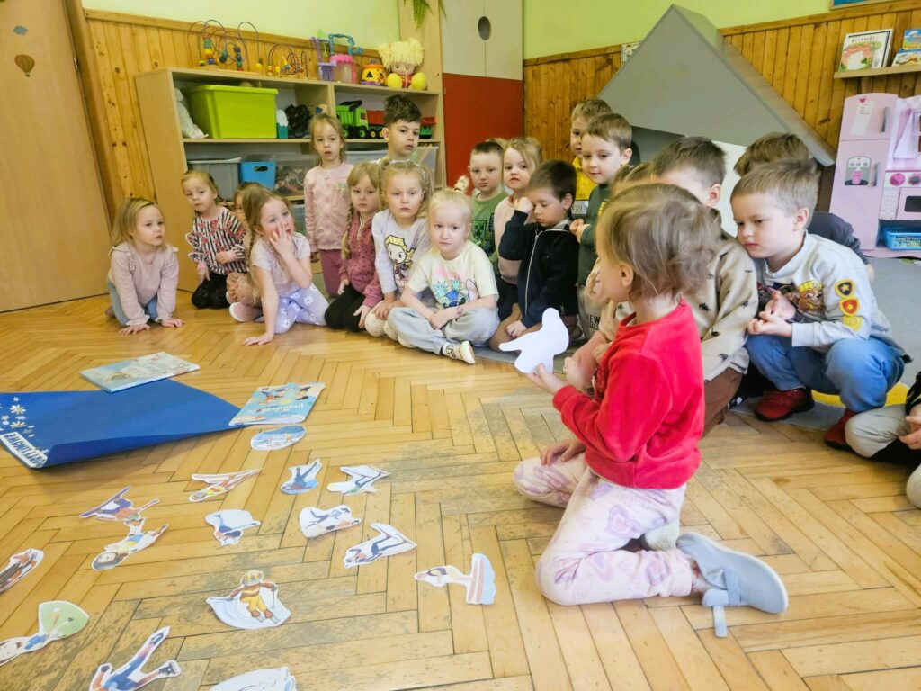 Na ziemi w sali bibliotecznej siedz przedszkolaki . jeden z nich rozwiązuje kalamury. Wszystkie dzieci ubrane kolorowo.
