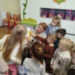 Pani bibliotekarka pokazuje grupie dzieci książkę . Za nimi kolorowe szafki.