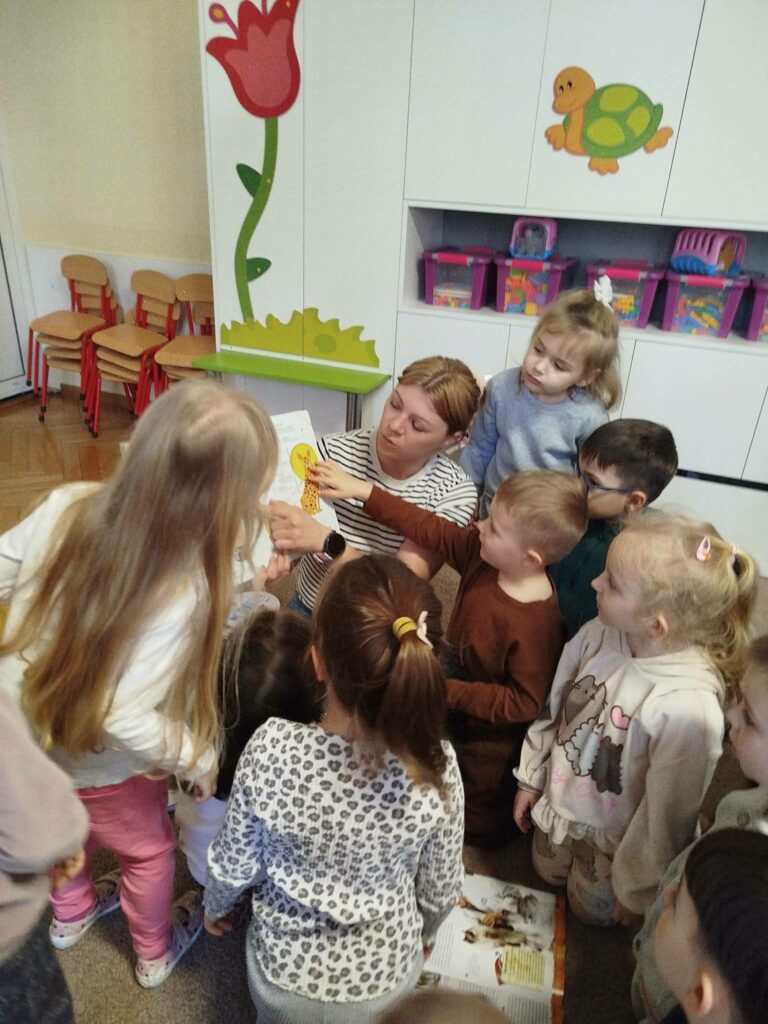 Pani bibliotekarka pokazuje grupie dzieci książkę . Za nimi kolorowe szafki.