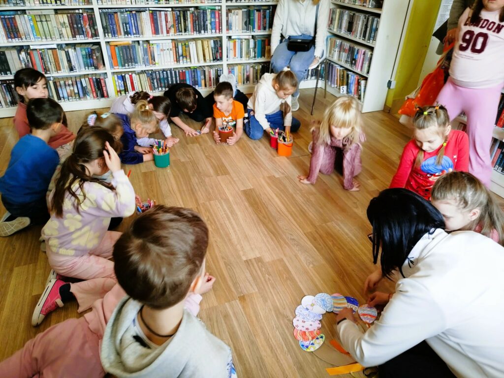 Na zdjęciu na ziemi w kółeczku siedzą dzieci i częśc z nich wykonuje prace plastyczne ,