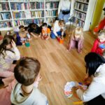 Na zdjęciu na ziemi w kółeczku siedzą dzieci i częśc z nich wykonuje prace plastyczne ,