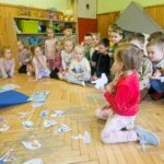 Na ziemi w sali bibliotecznej siedz przedszkolaki . jeden z nich rozwiązuje kalamury. Wszystkie dzieci ubrane kolorowo.