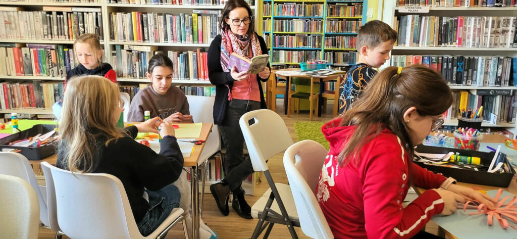 na zdjęciu w sali biblioteczne przy stołach siedza dzieci i wykonuja prace plastyczne wycinanki . Wokół nich stoi pani bibliotekarka i czyta im książkę