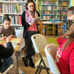 na zdjęciu w sali biblioteczne przy stołach siedza dzieci i wykonuja prace plastyczne wycinanki . Wokół nich stoi pani bibliotekarka i czyta im książkę