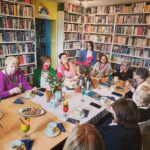 na zdjęciu widać grono kobiet siedzące wokół stołu w sali bibliotecznej. Na sole kawa w filiżankach, kwiaty, ciastka. Wokół regały z kolorowymi książkami.i