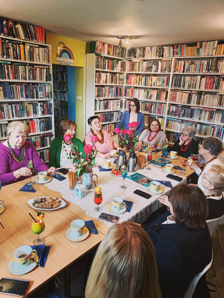 na zdjęciu widać grono kobiet siedzące wokół stołu w sali bibliotecznej. Na sole kawa w filiżankach, kwiaty, ciastka. Wokół regały z kolorowymi książkami.i