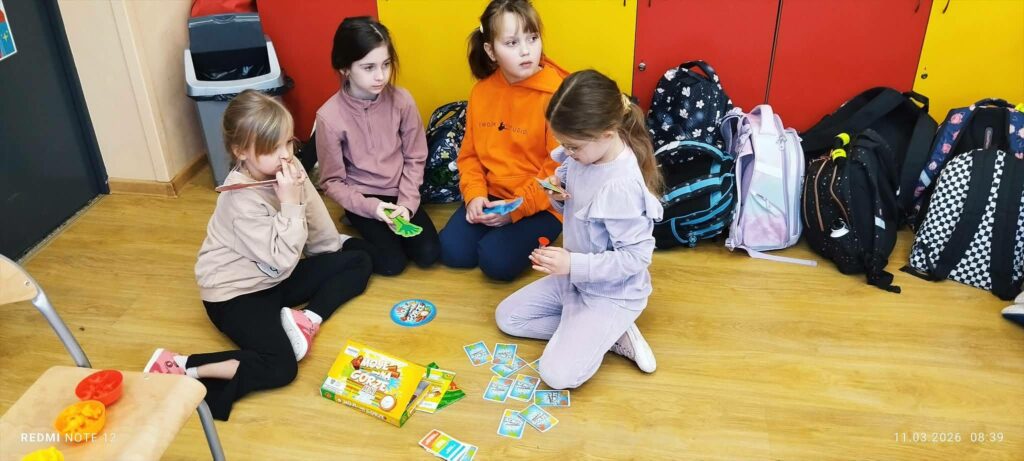 czery dziewczynki siedza na ziemi i graja w krę. Za nimi plecaki.