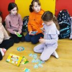 czery dziewczynki siedza na ziemi i graja w krę. Za nimi plecaki.