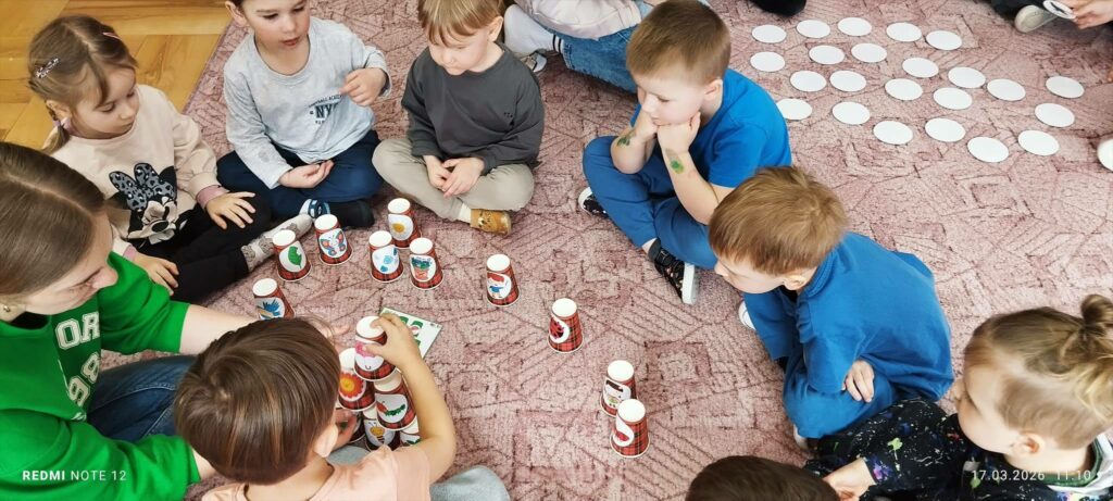 Ośmioro dzieci siedzi w kółku na podłodze. Przed nimi kubki poodwracane. Dzieci układają kubki