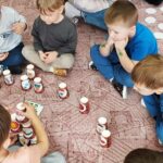 Ośmioro dzieci siedzi w kółku na podłodze. Przed nimi kubki poodwracane. Dzieci układają kubki
