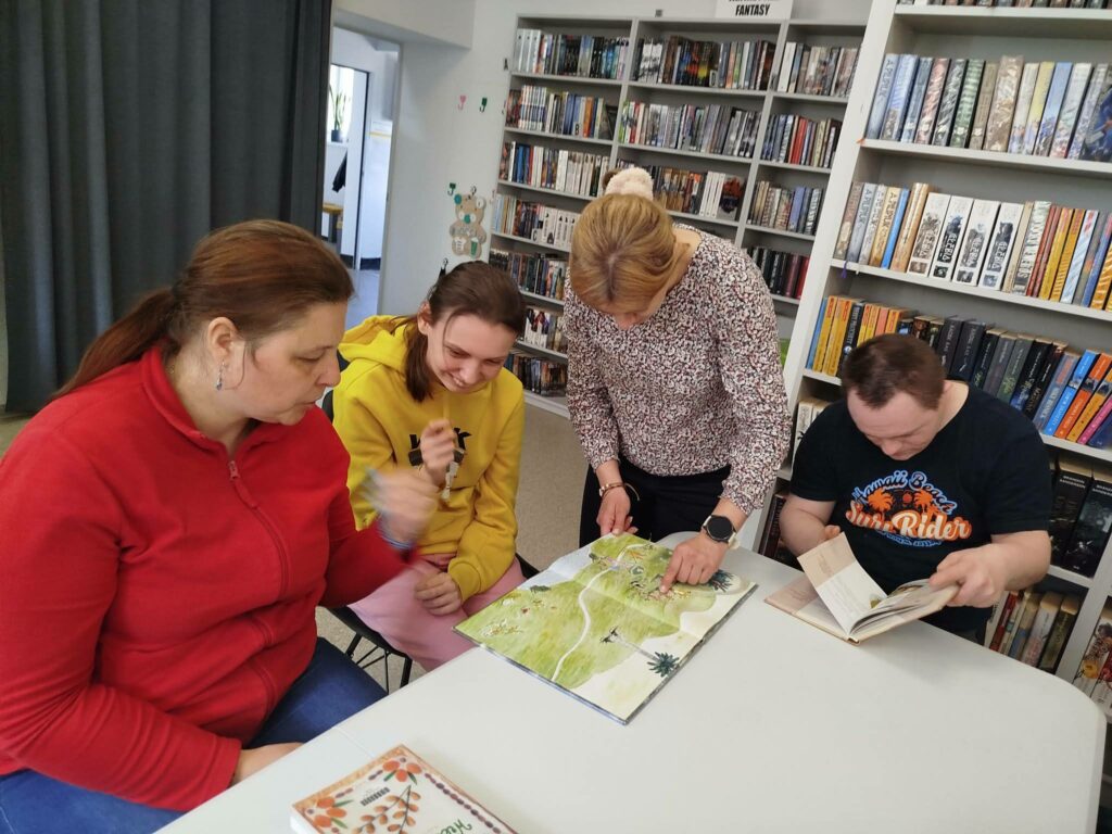 trzy osoby siedzą przy stole o oglądają ksiązki. Przed nimi stoi pani i prezentuje książkę.