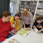 trzy osoby siedzą przy stole o oglądają ksiązki. Przed nimi stoi pani i prezentuje książkę.