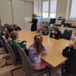 W sali bibliotecznej przy stole wokół siedzą dzieci i słuchaja pani, która prezentuje książkę.