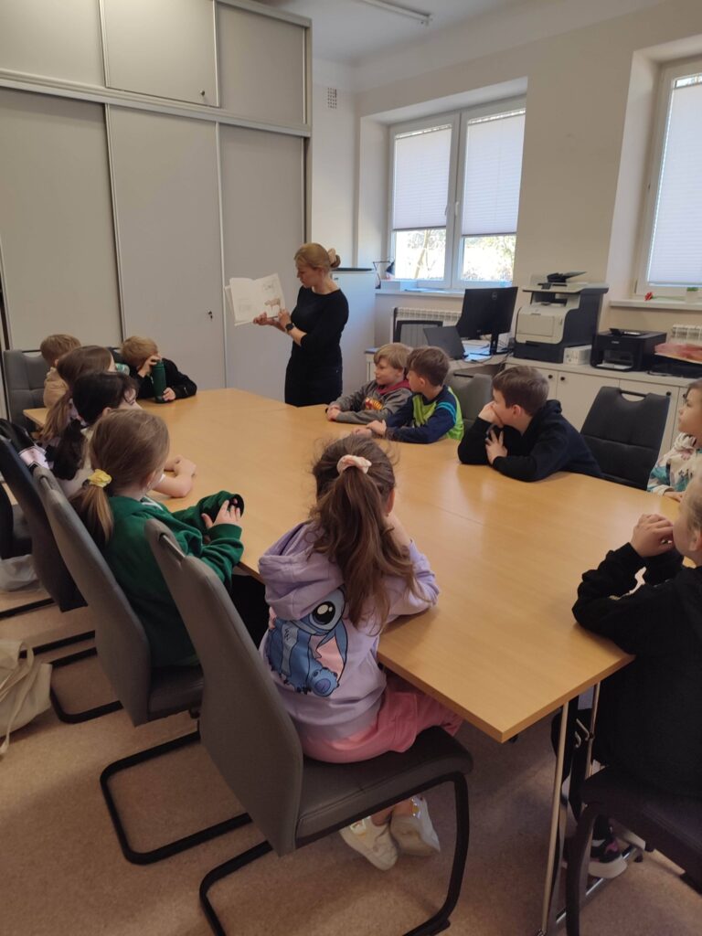 W sali bibliotecznej przy stole wokół siedzą dzieci i słuchaja pani, która prezentuje książkę.