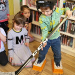 Na zdjęciu czwórka przedszkolaków. jeden z nich trzyma w ręce kij do hojeja i stoi w łyżwach zrobionych z tektury. na głowie ma kask . Dzieci przyglądają sie chłopcu.