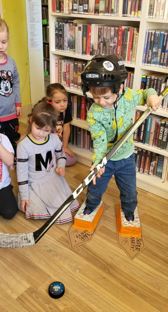 Na zdjęciu czwórka przedszkolaków. jeden z nich trzyma w ręce kij do hojeja i stoi w łyżwach zrobionych z tektury. na głowie ma kask . Dzieci przyglądają sie chłopcu.