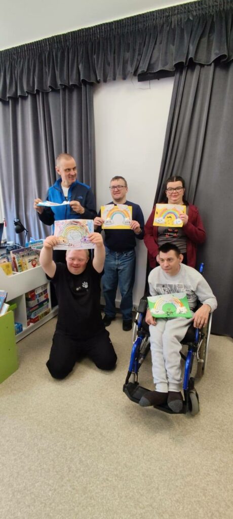 pięc osób na tle zasłony szarej prezentuje wykonane przez siebie prace. Jeden chłopiec siedzi na wózku inwalidzkim