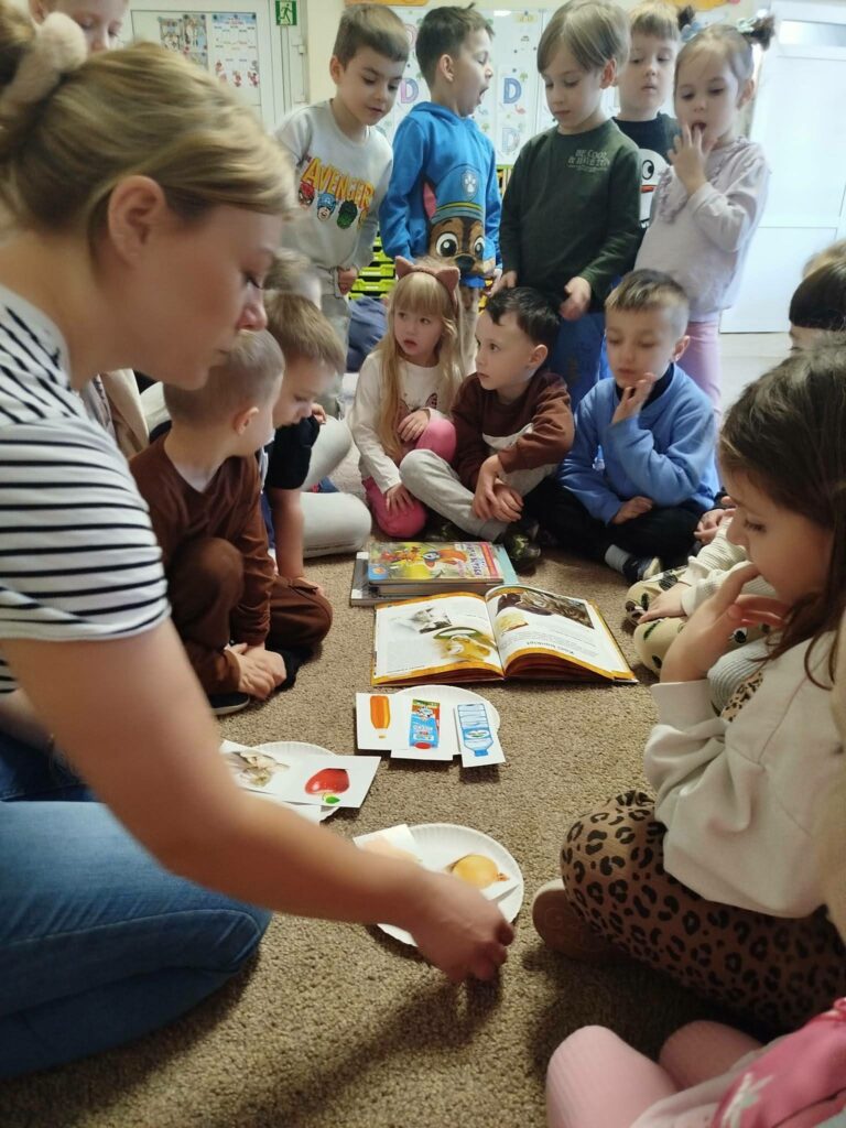 na zdjęciu pani bibliotekarka wraz z dziećmi siedzi na podłodze . Część dzieci stoi . Na ziemi porozkładane kartki z zagadkami i ksiązki