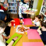 Na zdjęciudzieci siedza przy stole. Przed nimi olorowe kartki. pani bibliotekarka rozdaje dzieciom artykuły plastyczne do wykonywanych prac .