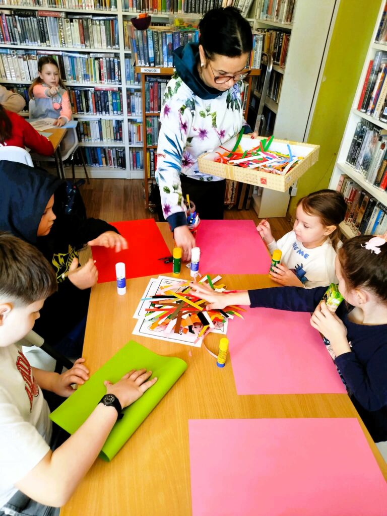 Na zdjęciudzieci siedza przy stole. Przed nimi olorowe kartki. pani bibliotekarka rozdaje dzieciom artykuły plastyczne do wykonywanych prac .