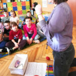 na zdjęciu widać kobietę klęczącą na podłodze i pokazująca dzieciom instrumenty