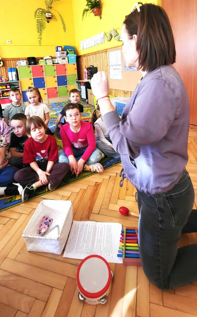 na zdjęciu widać kobietę klęczącą na podłodze i pokazująca dzieciom instrumenty