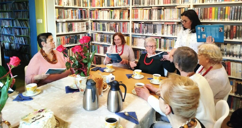na zdjęciu widać grono kobiet siedzące wokół stołu w sali bibliotecznej. Na sole kawa w filiżankach, kwiaty, ciastka. Wokół regały z kolorowymi książkami.i