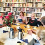 na zdjęciu widać grono kobiet siedzące wokół stołu w sali bibliotecznej. Na sole kawa w filiżankach, kwiaty, ciastka. Wokół regały z kolorowymi książkami.i