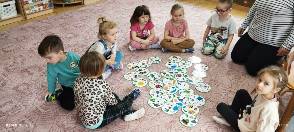 Dzieci siedzą na podłodze. w kółku. Przed nimi rozrzucone okrągłe karty tekturowe z ilustracjami.