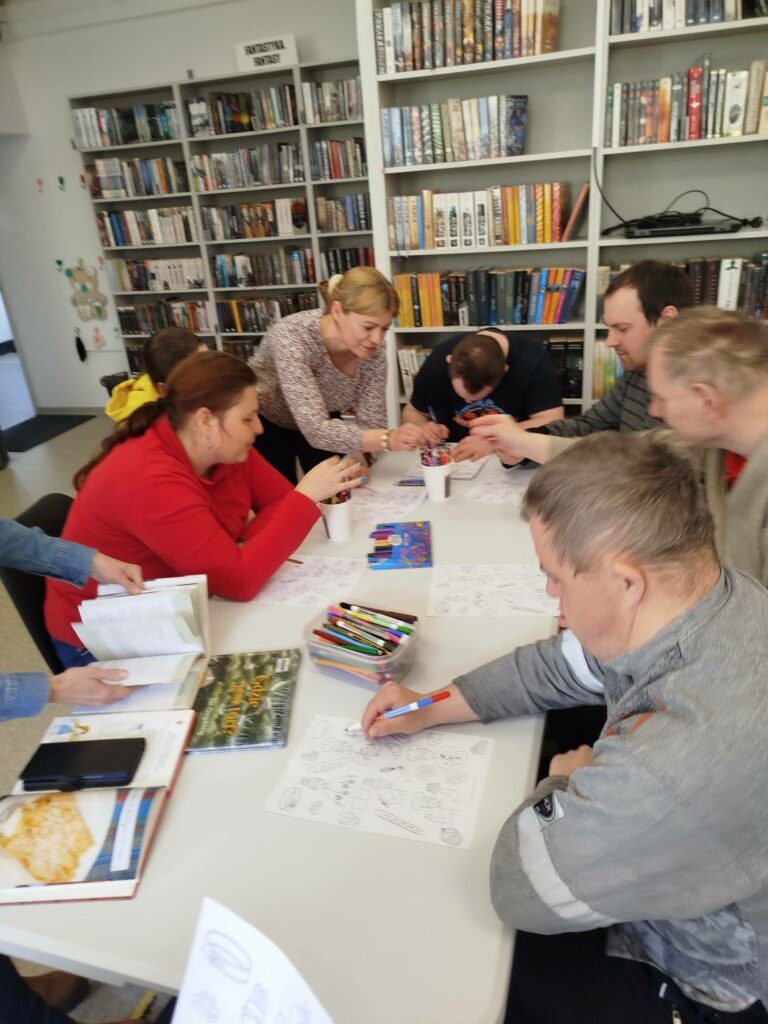 Przy stole siedem osób i wykonkja prace plastyczne.- kolorują. Na stole kredki, książki.