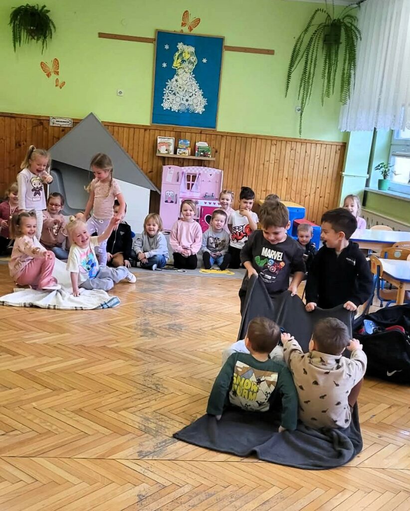 Na ziemi siedza dzieci w sali przedszkolnej. Czterech chłopców uczestniczy w dyscyplinie dwóch chłopców siedzi na kocyku i dwóch ich ciągnie .