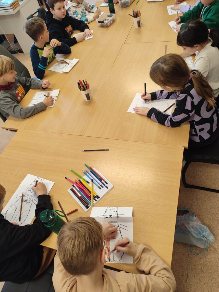 Ptzy stole wsali bibliotecznej siedza dzieci uczestniczce w lekcji bibliotecznej i wykonujące prace plastyczne Kapibarę