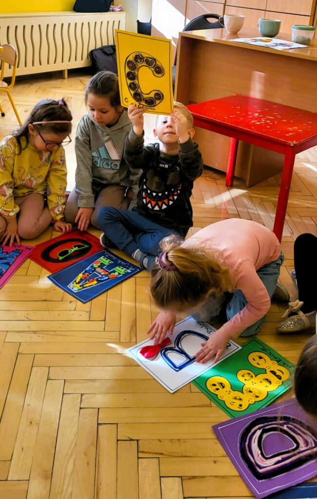na zdjęciu widać dzieci siedzące na podłodze i oglądające kolorowe literki