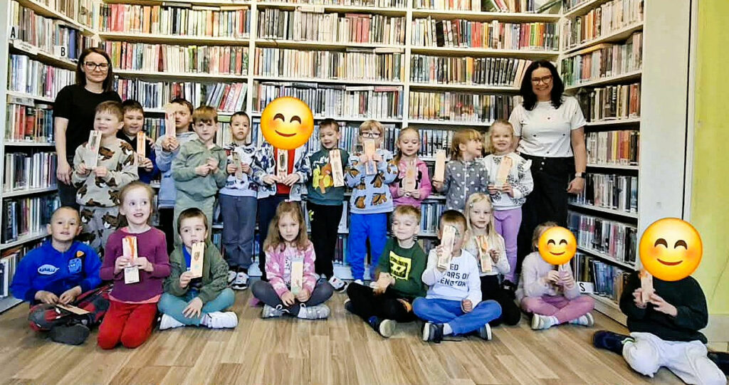 na zdjęciu widać dzieci stojące w sali bibliotecznej na tle regału z książkami