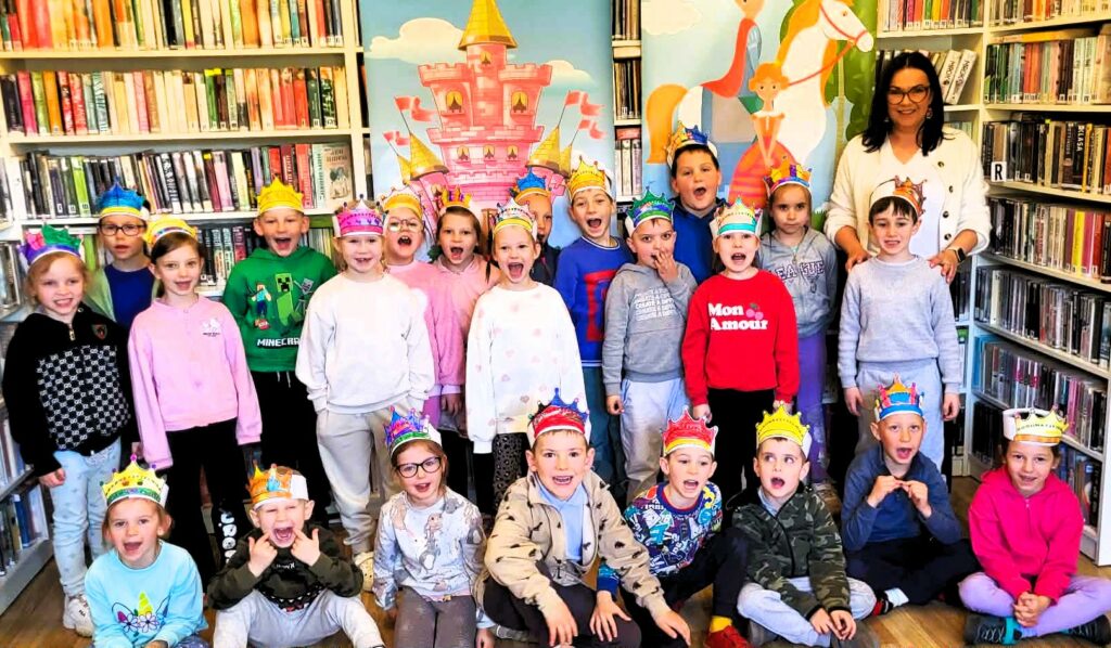 na zdjęciu widać dzieci stojące w sali bibliotecznej. Każde z dzieci ma na głowie papierową koronę.