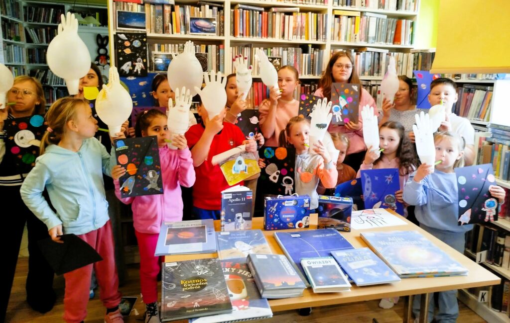 Na zdjęciu Dzieci stojące w sali bibliotecznej i trzymające kolorowe rysunki