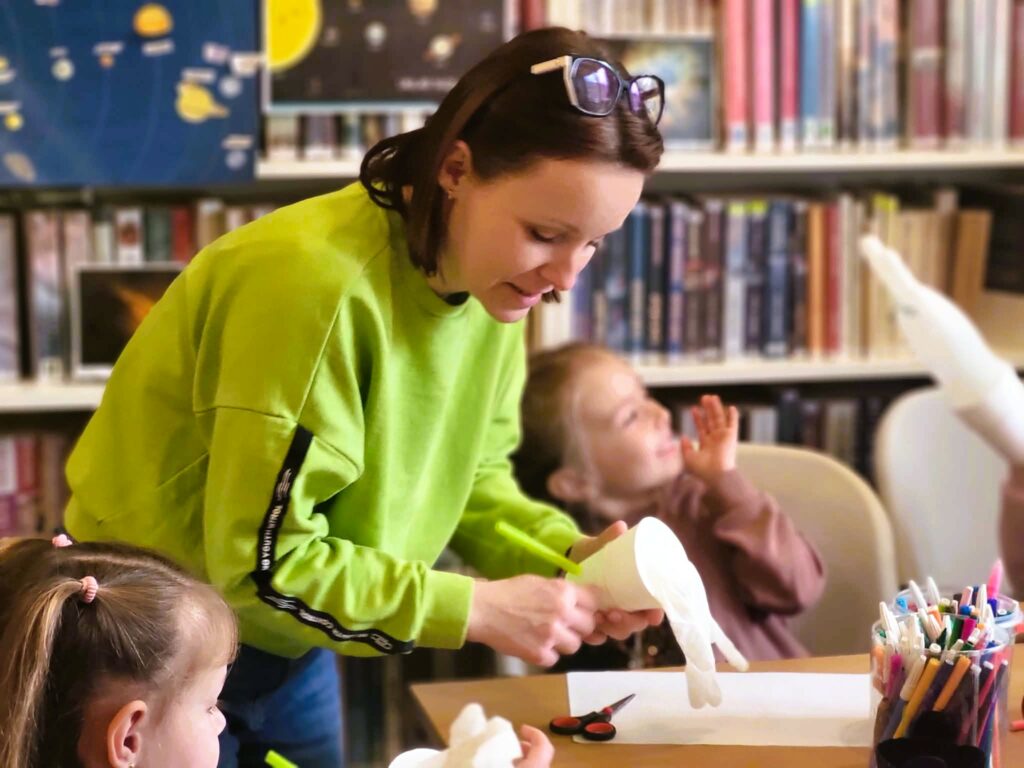 na zdjęciu widać bibliotekarkę pomagającą dzieciom w wykonywaniu pracy plastycznej
