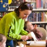 na zdjęciu widać bibliotekarkę pomagającą dzieciom w wykonywaniu pracy plastycznej