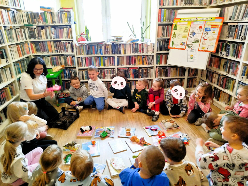 na zdjęciu widać dzieci siedzące na podłodze w sali bibliotecznej wokół książek