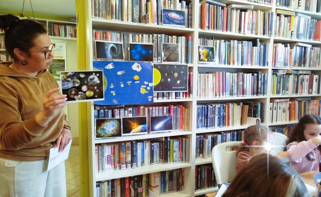 na zdjęciu widać bibliotekarkę pokazująca dzieciom zdjęcia przedstawiające kosmos