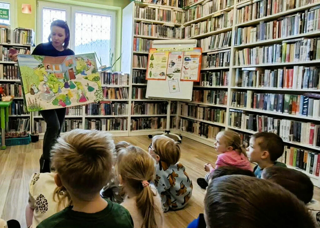 na zdjęciu widać kobietę pokazującą dzieciom dużą książkę