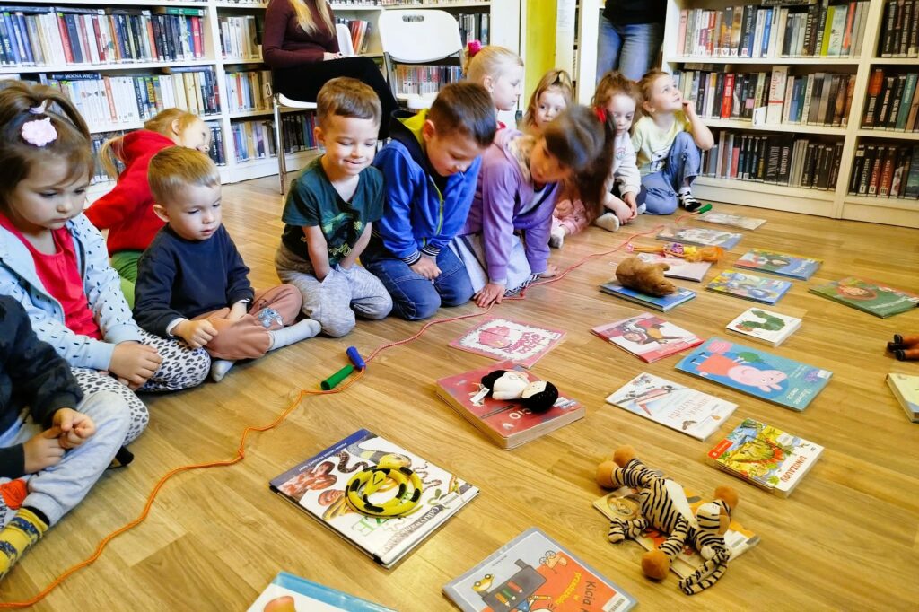 na zdjęciu widać dzieci na podłodze w sali bibliotecznej a przed nimi poukładane zabawki