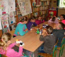 dzieci podczas wakacyjnych zajęć w Bibliotece z uwagą słuchają opowieści czytanej przez jedną z dziewczynek