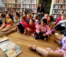 na zdjęciu widać dzieci w sali bibliotecznej, siedzące na podłodze a przed nimi książki